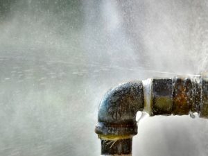 Close-up of leaking water pipes with water spraying from a corroded joint, illustrating the impact of corrosion.