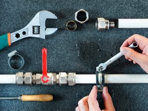 Hands repairing leaking water pipes using wrenches, with various plumbing tools and pipe fittings arranged on a work surface.