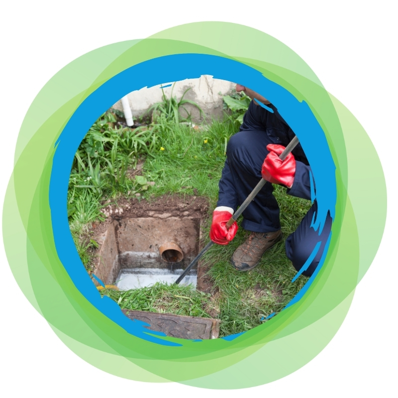 Technician clearing an outdoor blocked drain using a rod near a garden area.