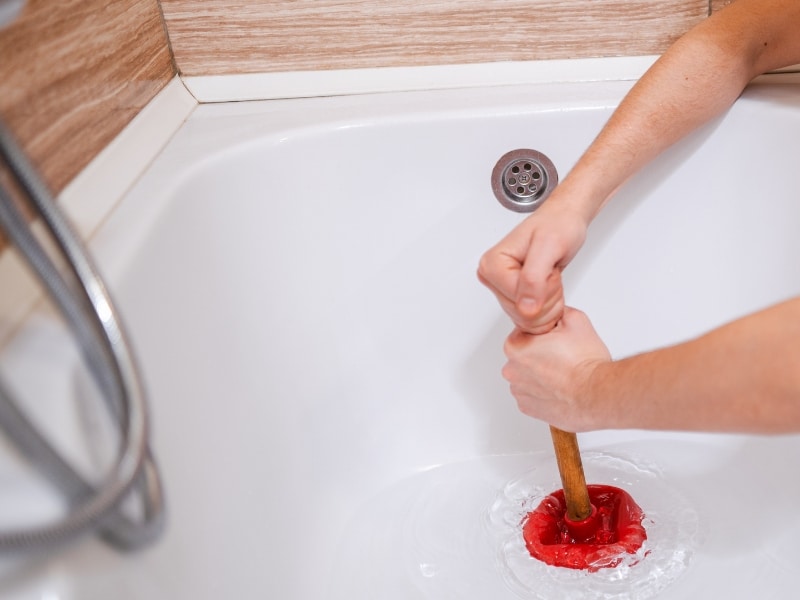 A person clears a bathtub blocked drain by plunging with a red rubber plunger.