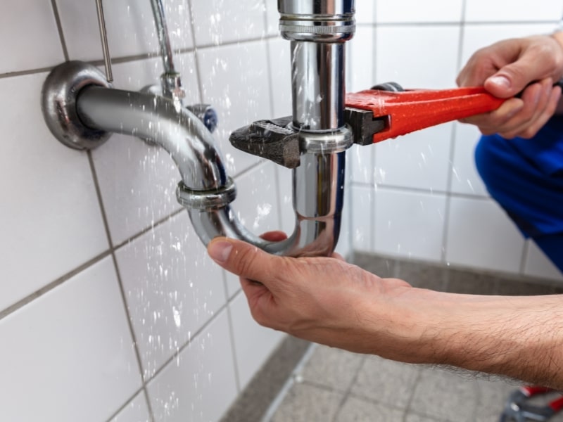 Plumber using red wrench to fix a leaking bathroom pipe spraying water under a sink