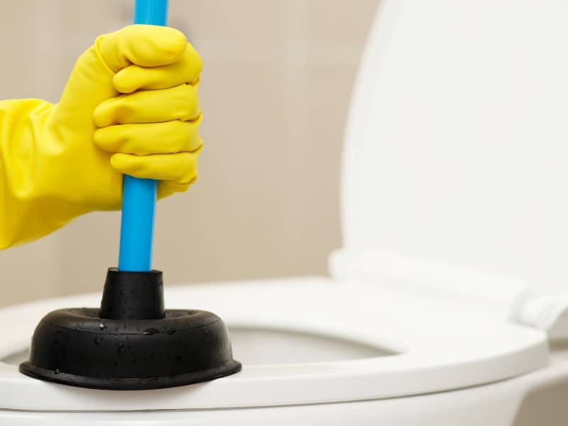 Close-up of gloved hand plunging a toilet blocked drain with a rubber plunger.

